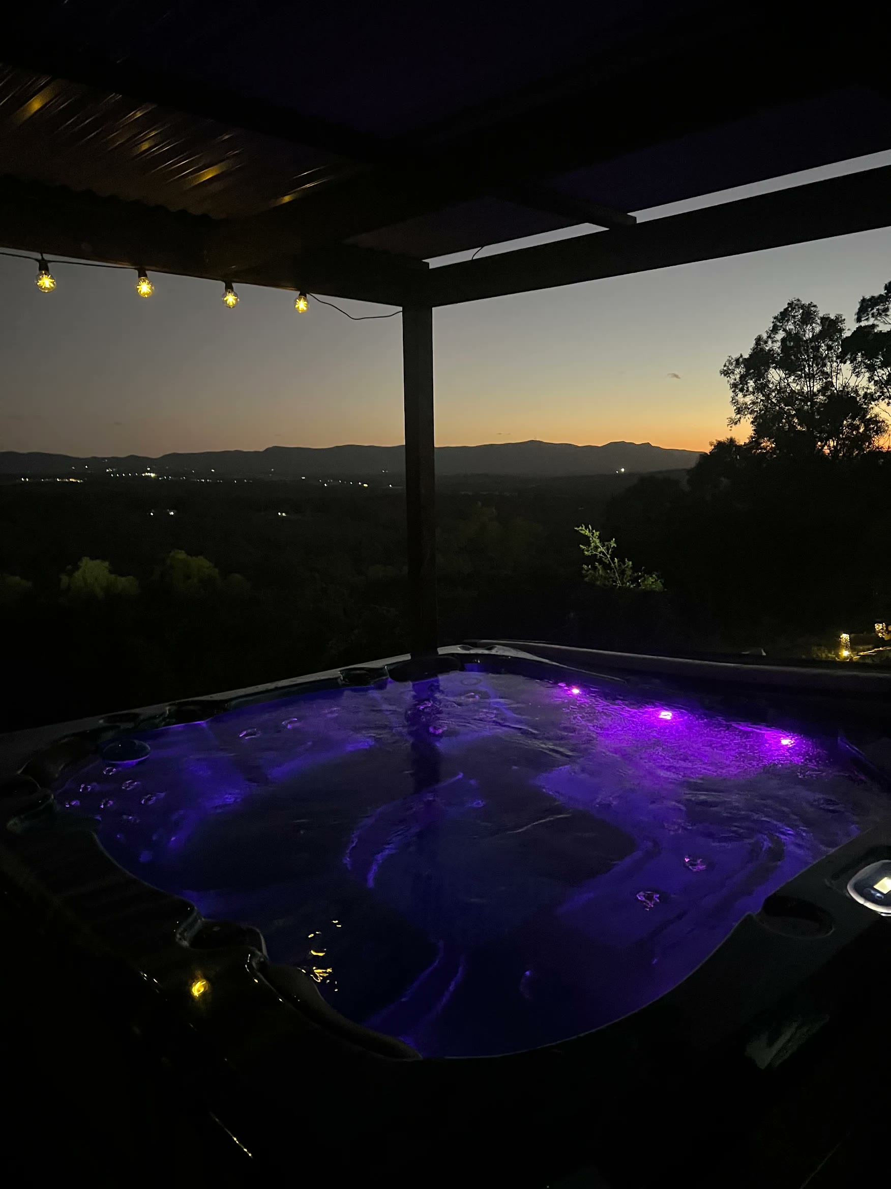 Purple-lit spa at dusk under a pergola with festoon lights, supplied by an Aqua Pulse customer